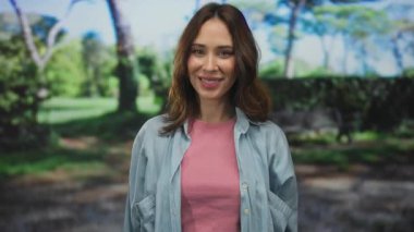 Pembe gömlekli ve kot ceketli mutlu İspanyol kadın güneşli bir parkta neşeli yeşil ağaçlar ve doğa ile çevrili.