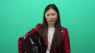 Kırmızı ceketli kadın yeşil arka planda miğfer tutuyor, güçlü bir ifade ve uzun saçlar sergiliyor, kendinden emin, şık bir hava yayıyor, modern, kentsel, kadın varlığını sembolize ediyor.