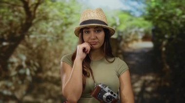 Parkta kameralı ve şapkalı İspanyol bir kadın güneşli bir günde dikkatlice ileriye bakıyor. Etrafı yemyeşil, açık hava macerasının özünü yakalıyor.