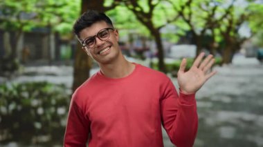 Gözlüklü genç adam gülümsüyor ve gün içinde ağaçlarla dolu canlı bir şehir sokağında el sallıyor..