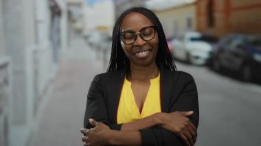 Gözlüklü bir kadın şehir caddesinde kendini kucaklıyor. Arabalar şehir yolunda sakin bir şekilde dolaşırken rahatlık ve yansıma yayıyor..