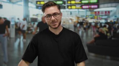 Sakallı, gülümseyen ve yukarıyı gösteren genç bir adam kalabalık bir havaalanı terminalinde duruyor. Arka planda insanlar var. Seyahat ve heyecan hissi taşıyor..