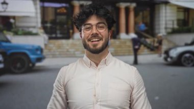 Sakallı İspanyol genç adam şehir caddesinde arabaların ve binaların bulanık geçmişiyle gülümserken şaşkınlık ve mutluluğu ifade ediyor..