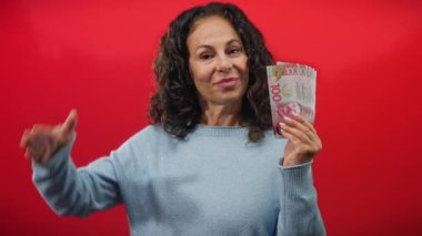 Soyutlanmış kırmızı arka plana karşı elinde yeni Zelanda doları tutan orta yaşlı bir kadın, canlı para birimi ve neşeli tavırlar sergiliyor, finansal refahı vurguluyor..
