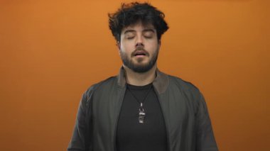 Latin kökenli genç adam gözleri kapalı meditasyon yapıyor ve turuncu arka plana karşı ellerini kaldırıyor. Rahatlama ve farkındalığı modern bir bağlamda aktarıyor..
