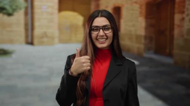 İspanyol bir kadın gülümsüyor ve eski bir üniversite kampüsünde kırmızı tişört ve gözlük takarak baş parmak işareti yapıyor..