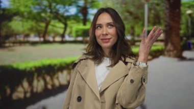 Parktaki bir kadın bej bir ceket giymiş havayı öpüyor. Doğanın yeşillikleriyle çevrili sakin bir an yaratıyor. Açık havada neşeli bir jest yapıyor..