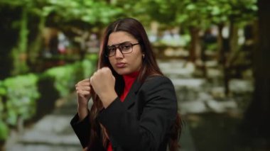 Yumruklarını sıkmış gözlüklü, kendine güveni tam bir İspanyol kadın, canlı bir park ortamında duruyor. Etrafı yemyeşil ve güneşli bir patikayla çevrili..