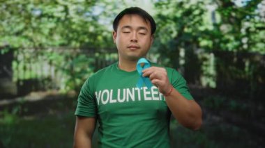 Gönüllü tişört giyen genç adam, kanser farkındalığı ve açık hava katılımını gösteren bir parkta mavi kanser kurdelesi tutuyor..