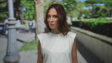 Beyaz tişörtlü genç İspanyol kadın canlı yeşil bir park ortamında tetikte bekliyor. Bulanık insanlar ve şehir geçmişi açık havada tetikte ve tetikte olmayı öneriyor..