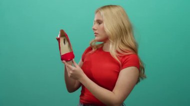 Sarı saçlı kadın yeşil arka planda kırmızı topuklu ayakkabı tutuyor. Moda tarzı ve zarafeti sergiliyor..