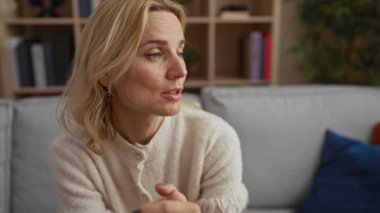 Rahat bir oturma odasında, gri bir kanepede konuşan bir kadın, sarı saçlar ve arka planda kitaplar ve bitkiler olan günlük giysiler.