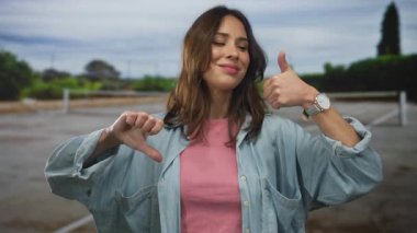 Gündelik kıyafetli genç bir kadın açık havada tenis kortunda baş parmak uçlarını yukarı aşağı sallayarak açık havada karışık duyguları gösteriyor..