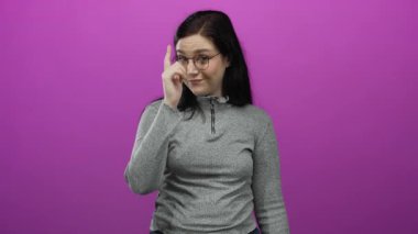 Gri süveter ve gözlüklü, canlı pembe arka planda, kendine güvenen bir ifadeyle doğrudan kameraya bakan kadın.