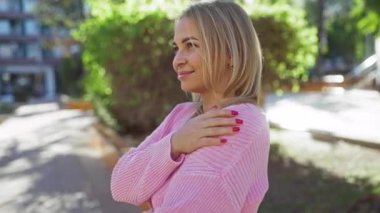 Pembe kazaklı kadın şehir parkında güneşli bir günün tadını çıkarıyor. Arka planda binalar, sıcak havalarda dışarıda huzur içinde gülümsüyor.