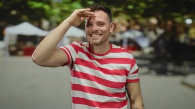Açık havada gülümseyen genç adam şehir sokaklarında arka planda bulanık, çizgili tişört giyen, şehir ortamında kendine güven ve pozitiflik gösteren baş parmağını kaldırıyor..