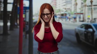 Kalabalık şehir sokaklarında kulaklarını tıkayan, bulanık şehir geçmişi olan, günlük kırmızı bluz giyen, stresli ve metropolitan ortamında arabalarla çevrili bir kadın..