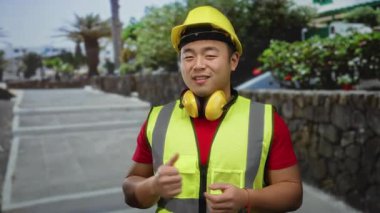 Sarı kasklı ve yelekli bir adam şehir sokağında bitkilerle dikiliyor, güneşli bir günde inşaat kıyafetleri ve aksesuarları sergiliyor..