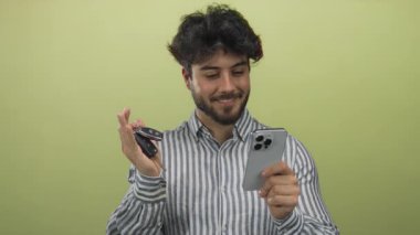 Sakallı ve çizgili tişörtlü genç adam elinde parlak sarı arka planda akıllı telefon ve araba anahtarlarıyla gülümsüyor..