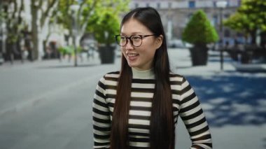 Gözlüklü ve çizgili süveter giyen gülümseyen kadın güneşli bir günde şehrin zarafetini resmediyor. Günlük tarzını ve doğal güzelliğini vurguluyor..