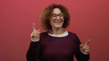 Orta yaşlı, esmer kıvırcık saçlı İspanyol kadın izole edilmiş kırmızı bir arka plan duvarında neşeyle dans ediyor, canlı ifade ve enerji yakalıyor..