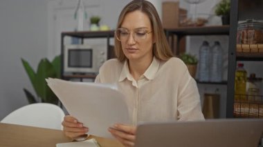 Parlak ofis ortamındaki belgeleri inceleyen kadın, iş yerinde dizüstü bilgisayar ve masa aksesuarlarıyla odaklanmış görünüyor. Bu da profesyonel bir ortam olduğunu gösteriyor..