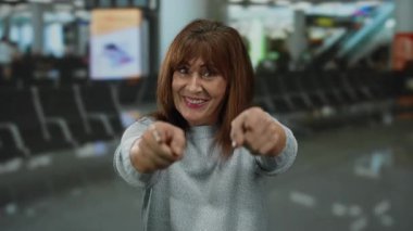 Gülümseyen bir kadın havaalanı terminalinde kollarını uzatarak arka planda tabela ve koltuklar ile dolu bir ev ortamında hoş geldin jesti yapıyor.