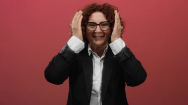 Orta yaşlı, esmer, kıvırcık saçlı İspanyol bir kadın yüzüne doğru ellerini indirirken gülümsüyor. İzole edilmiş kırmızı bir arka plan, neşe ve güven taşıyor..