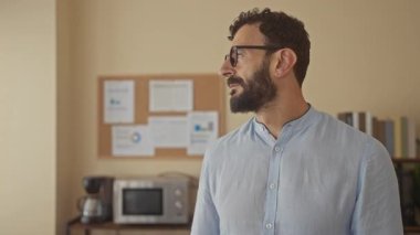 Gözlüklü ve sakallı bir adam düşünceli bir şekilde ofiste duruyor. Etrafı ilan panosu, mikrodalga fırın, kitaplar, tarafsız duvarlara dayanan kahve modern çalışma alanlarıyla çevrili..