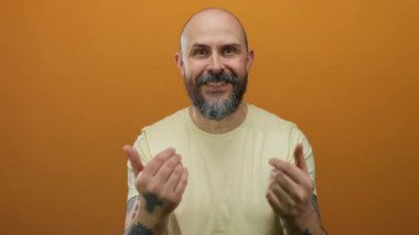 Kel sakallı adam, elleri turuncu duvara karşı hareketler yapıyor dövmeler ve davet edici ifadeler sergiliyor, izole bir stüdyoda sarı bir gömlek giyiyor..