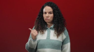 Kıvırcık saçlı kadın, kırmızı arka plana karşı kuşkulu bir ifade takınarak izole stüdyo ortamında şüphe ve belirsizliği işaret ediyor..