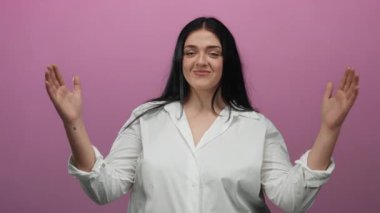 Büyük beden bir kadın, beyaz gömlekli, elleri pembe arka planda, kendine güveni ve zarafeti canlı bir ortamda somutlaştırıyor..