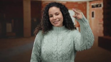 İnşaat alanında gülümseyen ve selam veren bir kadın. Samimi bir süveterin içinde selam veren bir jestle kendine güveni ve sıcaklığını gösteriyor..