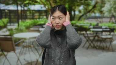 Terasta kulakları tıkayan bir kadın. Bulanık şehir kafesi, gri kazak ve pembe kollu Asyalı yetişkinleri şehir ortamında resmediyor..