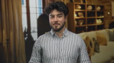 Otelde sakallı genç bir adam çizgili gömleğiyle rahat bir şekilde gülümsüyor rahat bir arka planda raflar ve mobilyalar sergiliyor.