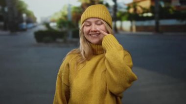 Sıcak sarı bir süveter ve bereyle buruna dokunan bir kadın, sakin bir şehir sokağında gün ışığında gülümsüyor, açık havada bir rahatlama ve sıcaklık anı yakalıyor..