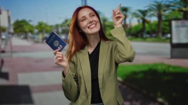 Dışarıda elinde Kore pasaportuyla gülümseyen, neşe saçan, kızıl saçlı, şehir ayarları seyahat hazırlığını ve pozitifliği belirten, arka planda bulanık sokak ortamı olan bir kadın..