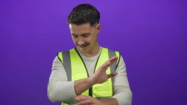 Yalıtılmış mor arka plan duvarının önünde elleri ile yansıtıcı yelek jestler giyen genç adam görünüşe göre oyuncu ya da küçümseyici bir şekilde.