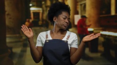 Önlük giymiş bir kadın kilisede yetişmiş, kapalı bir dini ortamda, bulanık figürler ve mumlarla çevrili bir şekilde tefekküre dalmış..