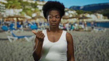 Sahilde arka planda deniz ve şemsiyelerle baş parmağını gösteren bir kadın, açık gökyüzü altında gevşek deniz havasını yakalıyor..