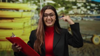 Genç İspanyol kadın deniz kıyısındaki bir yeri işaret ediyor. Elinde açık mavi gökyüzü ve uzak evleri olan güneşli bir arka planda kırmızı bir pano var..