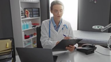Klinikte yazan kadın doktor gözlük ve steteskop takarak hastane çalışma alanında tıbbi ekipman ve belgelerle profesyonelliği gösteriyor..