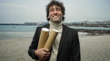 Sahilde iki silindirik objeyi tutarken güvenle gülümseyen bir adam, rahat bir balıkçı yaka kazağı ve ceket giymiş, cazibesini ve tarzını, sakin bir sahilin arka planına karşı sergiliyor..