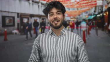 Sakallı genç adam kalabalık bir caddede duruyor, canlı bir açık hava ortamında şehir yaşamını sergiliyor ve arka planda çeşitli insanlarla şenlikli dekorasyonlar yapıyor..