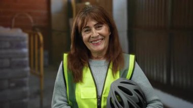 İçeride inşaat alanında gülümseyen, güvenlik yeleği giyen ve arka planda endüstriyel malzeme ve aletlere karşı kask tutan İspanyol kıdemli kadın..