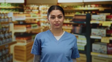 Mavi önlük giyen kadın, çeşitli ürünlerle dolu bir süpermarket koridorunda, yoğun bir mağaza ortamında duygularının bir karışımını taşıyor..