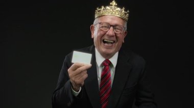 Takım elbiseli kıdemli işadamı, elinde kredi kartı tutarken gülüyor, siyah arka planda izole edilmiş, mali başarıyı ve liderliği simgeliyor..