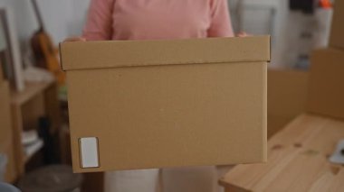 Yeni bir eve taşınırken bir kadının oturma odasında kutu paketlemesi orta yaşlı bir bireyin yeni bir yerleşim yerine geçişini yansıtıyor..