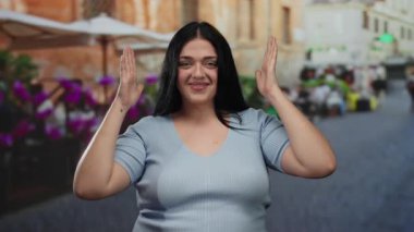 Canlı bir şehir caddesinde neşeli bir şekilde el kol hareketi yapan kadın, şehir manzarasının ortasında sıcak bir gülümsemeyle vücut özgüveni ve pozitifliğini gösteriyor..