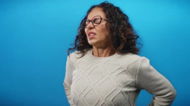 Acı çeken, gözlük takan, süveter giyen, parlak mavi arka planda duran, rahatsızlık ve olgunluğu ifade eden kıvırcık saçlı ve İspanyol özellikli bir kadın..